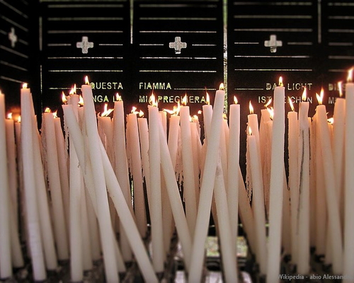 Viele brennende Kerzen in einer Votivkerzenwand in Lourdes, im Hintergrund Schrifttafeln mit dem Text „Diese Flamme ist mein Gebet“ in mehreren Sprachen.