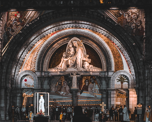 Gläubige versammeln sich am kunstvoll beleuchteten Portal der Basilika von Lourdes bei einer abendlichen Zeremonie mit Marienstatue, Kreuz und Prozessionsteilnehmern.