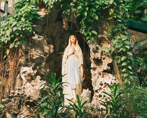 Statue der Jungfrau Maria in einer mit Pflanzen bewachsenen Grotte aus Naturstein, umgeben von tropischem Grün und Blumen.