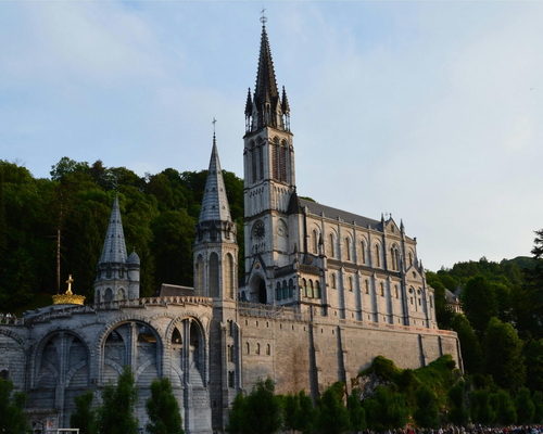 Seitenansicht der Basilika der Unbefleckten Empfängnis in Lourdes mit ihren markanten Türmen, umgeben von grünen Hügeln und Wäldern bei Abendlicht.