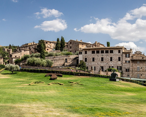 Historische Steinhäuser in Assisi oberhalb einer gepflegten Grünfläche mit Buchsbaumornamenten, unter leicht bewölktem Himmel.