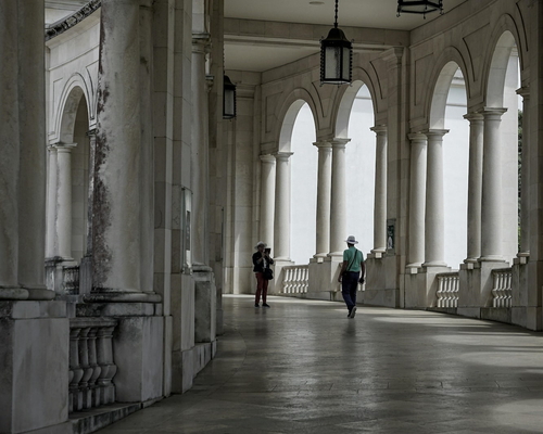 Überdachter Säulengang mit Rundbögen im Heiligtum von Fátima, zwei Personen gehen in Richtung des Ausgangs, Lampen hängen von der Decke.